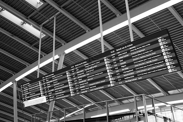 Utrecht, The Netherlands. February 19th, 2026: Black and white photograph of a digital departure board displaying train schedules inside Utrecht Central railway station. The modern station architecture with steel beams and glass elements frames the information display, symbolizing public transport, travel planning, infrastructure, and urban mobility.