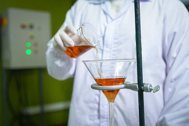 A technician in a white lab coat and gloves pours amber-colored natural extract—made from botanical ingredients and honey—into a glass funnel for filtration, with sample bottles and test tubes on the bench, representing natural product processing, formulation, and quality control.