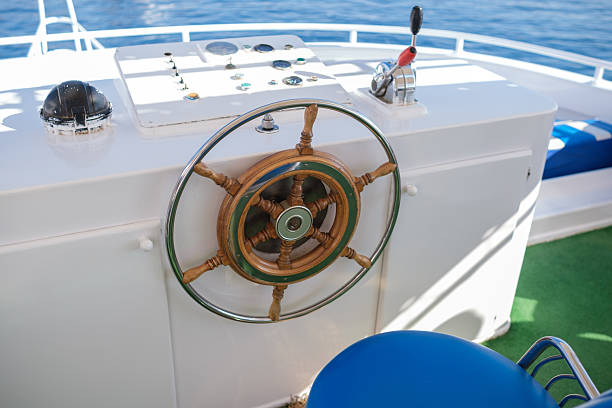 Boat steering wheel features a wooden rim and control panel on deck, with a blue seat and ocean water visible in the background
