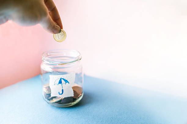 Hand dropping euro coin into glass jar labeled with umbrella symbol, representing building a rainy day fund and financial safety net.