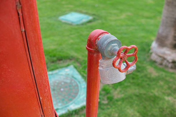 Red fire hydrant valve features a flower-shaped handle, situated on a grassy surface with utility covers seen in the background