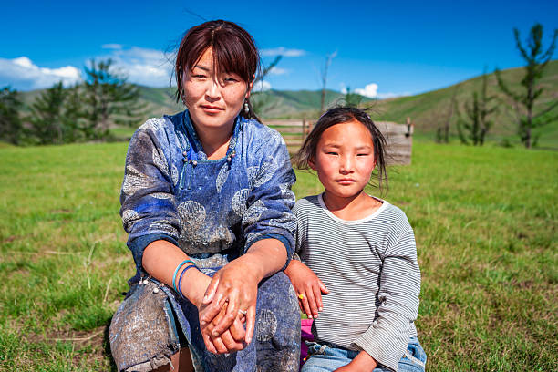An adult woman and a young girl sit close together on a sunlit grassy plain beneath a vivid blue sky. The woman wears a blue, patterned garment with beaded details and bracelets, her hands clasped in front of her; the girl wears a striped long-sleeve shirt and jeans, both with wind-tousled hair and slightly flushed, sun-kissed faces. Rolling green hills, scattered trees and a simple wooden fence form a calm rural backdrop.