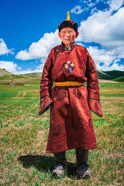 Full-body portrait of an elderly man standing on a green grassy steppe beneath a bright blue sky dotted with white clouds. He wears a richly patterned rust-red traditional robe with a yellow sash, a pointed black-and-gold hat, and well-worn leather boots. Small pins or medals are visible on his chest. The image emphasizes his weathered face and dignified posture against rolling hills and open landscape, conveying cultural heritage and rural life.