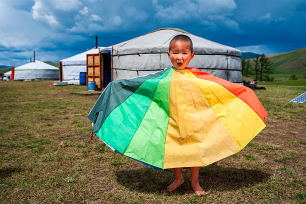 A young child stands barefoot on a grassy plain in front of white gers (yurts), draped in or holding a large rainbow-colored fabric that fans out like a cape. The child has an open-mouthed, surprised expression. Low, dramatic storm clouds fill the blue sky above, and open tent doors, barrels, and rolling green hills are visible in the background, suggesting a temporary camp on the steppe.