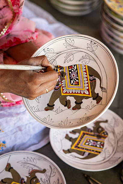 A close-up image of an artisan's hand delicately painting a decorative ceramic plate featuring a stylized elephant adorned with an ornate saddle cloth. The artist uses a fine brush to apply bright geometric and floral patterns in yellow, purple, red and black while surrounding foliage outlines remain lightly sketched. Several similar plates sit blurred in the background, highlighting the repetitive, detailed handcrafting process and the textured surface of the paint.