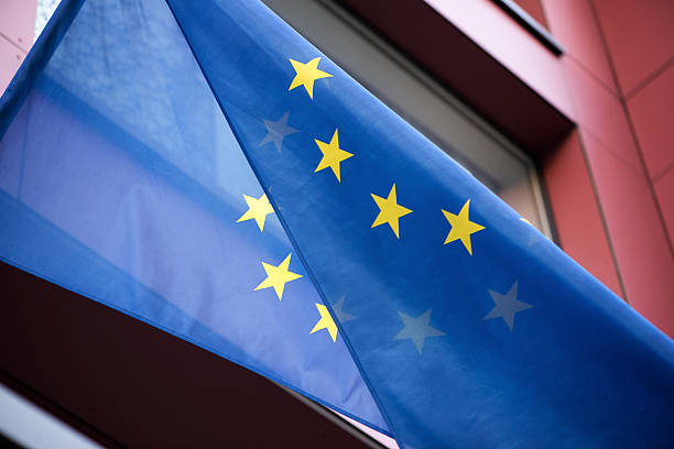 Vertical image showing a blue European Union flag with yellow stars hanging diagonally in front of a red modern building facade. Clean political symbol with strong color contrast.