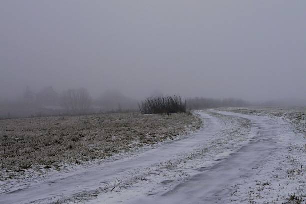 A wide-angle winter shot of a rural dirt road covered in snow and ice, disappearing into a thick, grey fog. The landscape is desolate and quiet, featuring frost-covered grass and a vast, hazy horizon. The minimalist composition and large overcast sky provide ample copy space, making it ideal for winter-themed backgrounds, travel concepts, or atmospheric editorial designs.