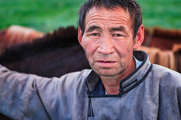 Close-up portrait of an older man with weathered skin, short dark hair and light stubble, wearing a grey, robe-style jacket. Deep lines and a thoughtful, slightly tired expression mark his face. He rests an arm on a blurred horse behind him; the background shows soft-focus green grass and the warm tones of animal coats.