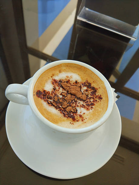 White cup and saucer of cappuccino coffee with chocolate on a glass table with sky reflection
