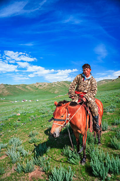 A lone rider sits astride a chestnut horse on a wide green steppe beneath a vivid blue sky filled with scattered clouds. He wears a richly patterned traditional coat and leather boots, holding simple reins as the horse grazes on grass and wildflowers. Low rolling hills and a few white gers (yurts) dot the distant landscape, creating a tranquil pastoral scene that evokes nomadic life and open-air freedom.