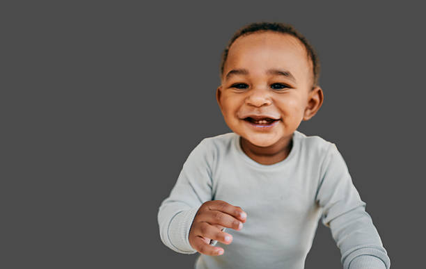 Young black baby smiling and reaching forward wearing a white shirt against a plain grey background with soft balanced lighting highlighting joyful expression