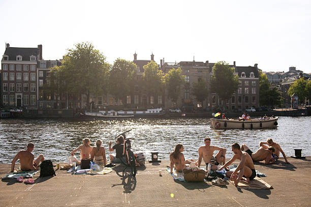 People enjoy the summer sun beside the Amstel River across from the H'ART Museum in Amsterdam, Netherlands (August 11, 2025)
