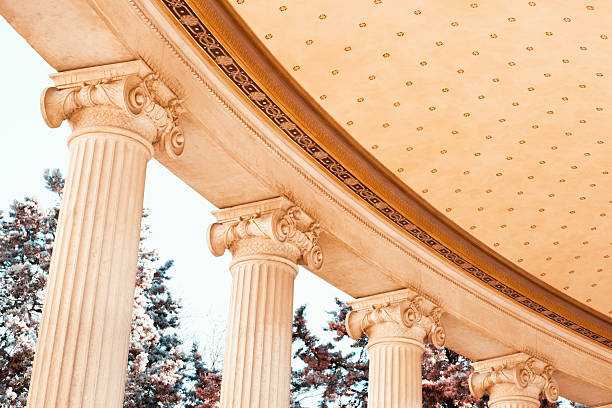 Close-up view of classical Ionic columns supporting a curved portico in Marseille. Decorative capitals and detailed ceiling patterns are illuminated by warm daylight. Soft blue sky and trees in the background contrast with the pale stone architecture, emphasizing texture and symmetry.