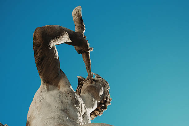 Close-up of a classical stone statue against a clear blue sky. The low-angle perspective emphasizes the sculpted details and dynamic pose of the figure. Strong natural light enhances the texture of the stone and the contrast with the background.