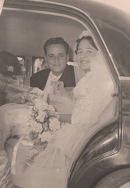 Image taken in the 1950s: happy couple posing after they goy married looking at camera sitting in a car