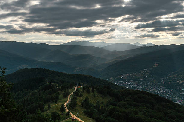 A dramatic view of a mountain valley with a winding dirt road leading through green hills and forested slopes. Sun rays break through heavy storm clouds, illuminating parts of the landscape and a small rural settlement below.