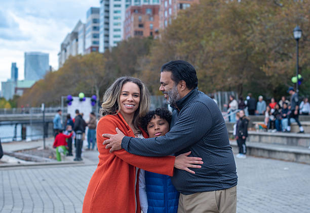 A multiracial family of three shares a group hug on a paved waterfront promenade. A mixed-race woman smiles at the camera while her Indian husband smiles at their son. Their elementary-aged boy is between them in the embrace. The family is enjoying an autumn afternoon outdoors in a park in Hoboken, New Jersey. City buildings and trees with fall colors are visible in the defocused background. This is a medium shot with an eye-level view and selective focus on the family.