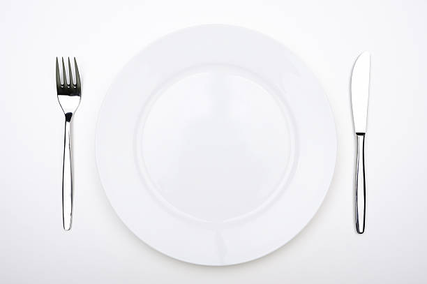 A fork and knife sit on each side of an empty white plate on a flat surface. The setup suggests it is time for a meal. The plate is clear of any food.