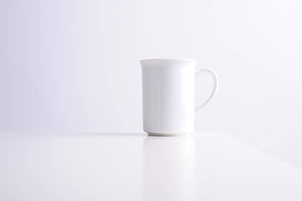 A plain white ceramic mug sits alone on a clean table. The background is light in color, and there are no other objects nearby.