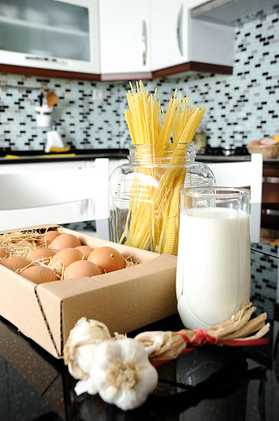 Items are arranged on a kitchen counter. A box holds eggs, a glass displays milk, and a jar contains dry pasta.