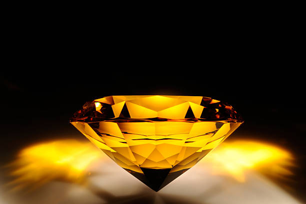 A yellow diamond is displayed at an angle, showing its facets. Light reflects off its surface and creates a glowing effect against a dark background.
