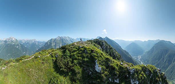 Hiker standing on a green mountain ridge in the Julian Alps, Slovenia, overlooking layered alpine valleys and distant peaks.