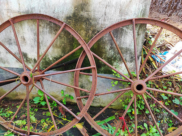 Two rusty, abandoned wagon wheels lean against a weathered concrete wall. It represents history, rural life, and industrial heritage, ideal for concepts of ageing, time, neglect, and sustainability.