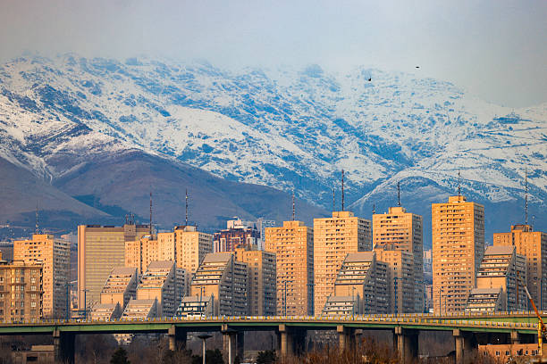 Urban skyline of Tehran with modern residential buildings and snow covered Alborz mountains in the background during golden hour. Atmospheric cityscape with warm evening light and winter landscape.
