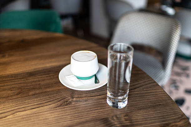 White cup of cappuccino with foamed milk and a glass of water on a natural wooden table, evoking a calm morning cafe scene with soft daylight and minimal, modern styling