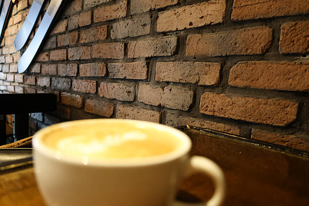 White coffee mug on wooden table in cozy cafe interior featuring exposed brick wall with vintage skis mounted as decoration. Rustic coffee shop atmosphere with authentic industrial design elements. Perfect for cafe culture, coffee lifestyle, small business, hospitality industry, breakfast concepts, and urban dining marketing content