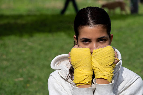 Athletic woman with yellow hand wraps looking at camera, ready for combat sport training in a park. Presenting determination and strength