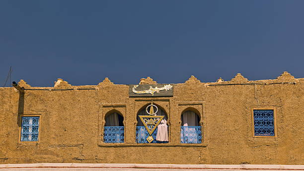 Rissani, Morocco-April 4, 2023: Barred and half-barred windows with arabesque grilles on the adobe facade of a traditional Berber house in the maydan or square facing the Moulay Ali Cherif mausoleum.