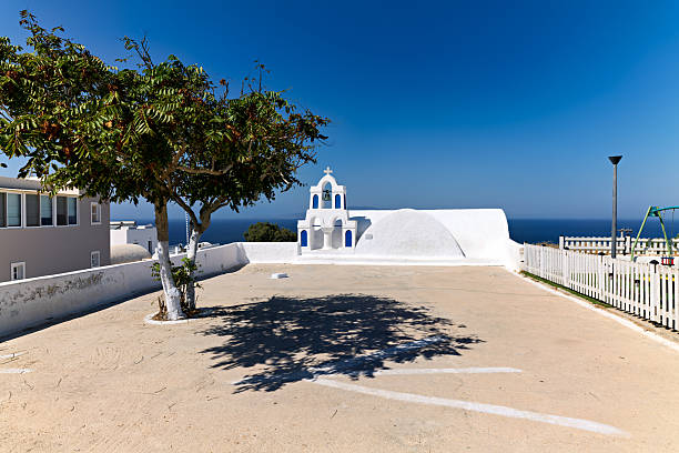 A classic white and blue Greek church with a bell tower, framed by a tree, overlooks the Aegean Sea on a sunny day in Santorini, Greece. Capturing island beauty.