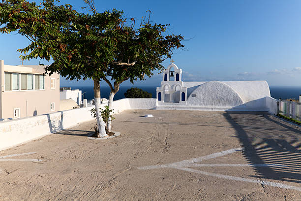 A traditional white church with a blue bell tower in Santorini, Greece, overlooks the Aegean Sea. A tree stands in the foreground, capturing the island's serene beauty.