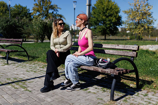 Two women are chatting while sitting on a wooden bench in a park. It is sunny, and the trees provide a natural background. They appear engaged in conversation and relaxed.