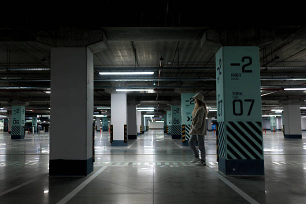 A woman is walking in a large parking garage with empty spaces around her. The environment is quiet and dimly lit, with visible concrete pillars and signs indicating levels.