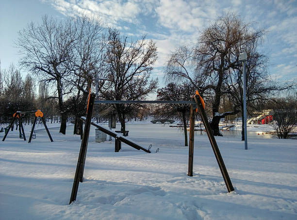 An empty playground blanketed in fresh snow featuring metal swing frames, bare trees and a lamp post in a quiet winter park. Soft afternoon light and blue sky create a calm, cold atmosphere ideal for seasonal, nature or urban recreation themes and for illustrating solitude and stillness.