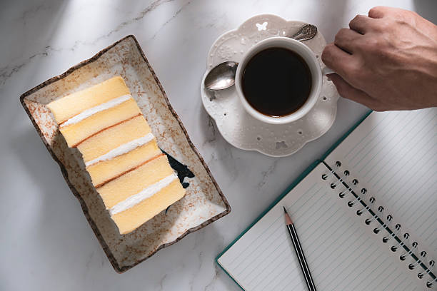 Hand of the man writing the book holds a coffee cup. With cake on marble background with soft natural sunlight.