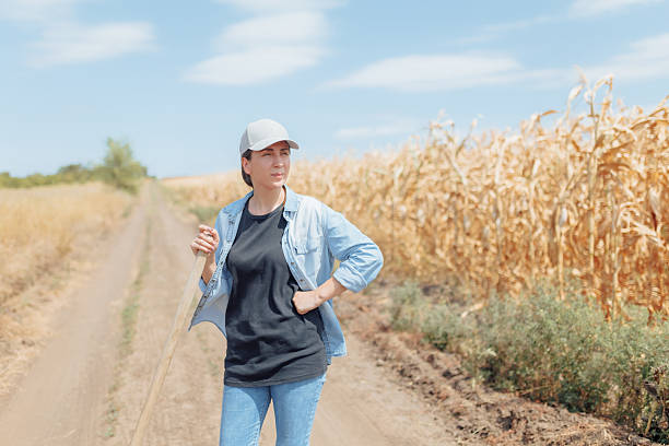 A smiling young woman in a cap and denim shirt stands in a sun-drenched cornfield, the bright sunlight casting a warm glow on her face.