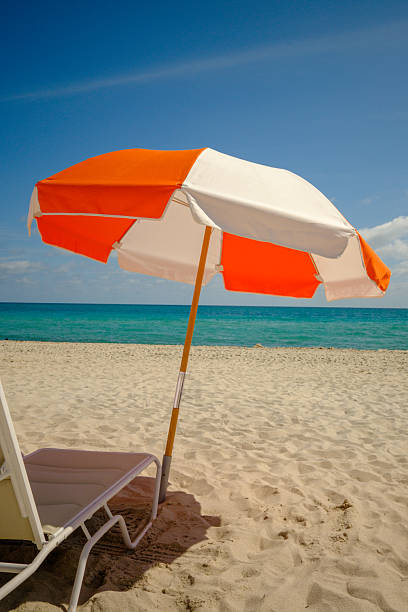 An orange and white umbrella and chaise lounge stand in the morning sun ready for beachgoers