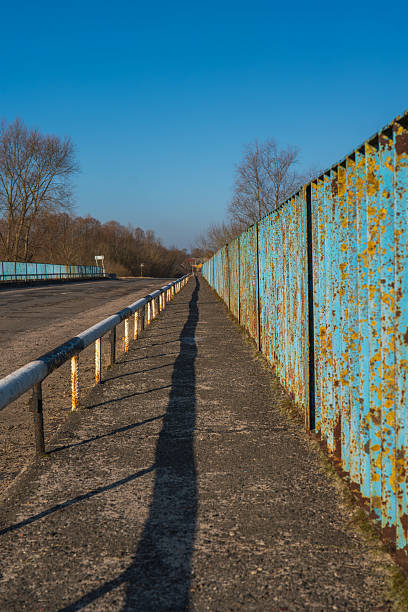 A narrow walkway runs beside a rusted turquoise metal fence, strong lines and harsh side light lead to a vanishing point, empty road and leafless trees suggest late autumn.
