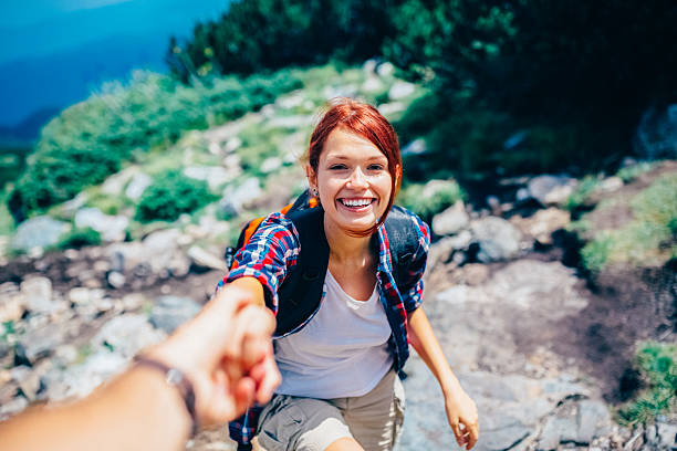 Close-up of a happy young woman hiker reaching forward and holding boyfriend hand as they help her climb on a mountain trail, with backpack and rocky landscape in the background. Outdoor adventure, teamwork, support, and travel lifestyle concept with dynamic perspective