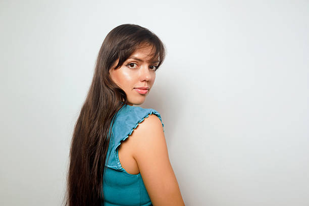 A woman stands with her long hair draping over her shoulder. She looks back with a slight smile in a simple room with light walls and natural light coming in during daytime hours.