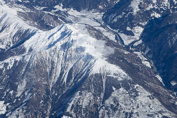 Bird’s-eye view of a snow-covered mountain ridge overlooking a winding valley dotted with small villages and frozen fields. Soft clouds drift across the peaks, while sunlight reveals the texture of the terrain and the vastness of the Austrian alpine region.