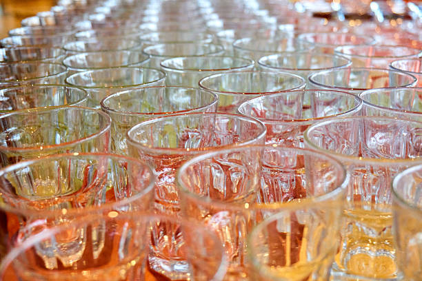 A close-up, high-angle shot of numerous empty glass tumblers arranged in neat rows, creating a sense of order and readiness. The glasses are clean and clear, reflecting ambient light with warm reddish-orange and yellow hues from their surroundings, likely a tabletop or serving surface. The depth of field is shallow, with the glasses in the foreground sharply focused while those receding into the background gradually blur, adding to the visual appeal and creating a sense of infinite repetition. The pattern of the glasses suggests preparation for an event, catering, or a large gathering.