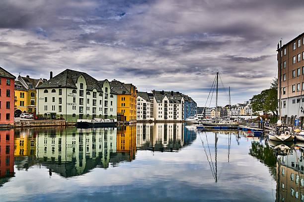 A stunning panoramic view of Ålesund, Norway, highlighting its iconic Art Nouveau (Jugendstil) architecture along the canal. Colorful buildings, including the prominent Hotel Brosundet, are perfectly mirrored in the calm waters, creating a symmetrical reflection. Sailboats and other vessels are moored in the picturesque harbor, framed by a dramatic, cloudy sky that adds a moody atmosphere to this beautiful Scandinavian cityscape.