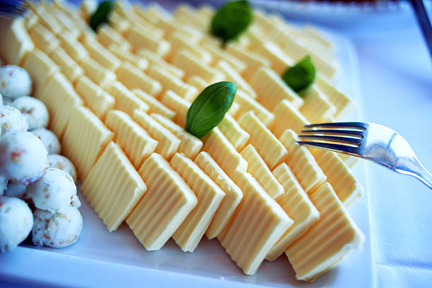 A vibrant close-up of a white serving platter featuring an elegant assortment of butter. Neatly arranged in diagonal rows are numerous light yellow butter slices, each with distinct ridged patterns, garnished with fresh green basil leaves. To the left, a pile of small, spherical herb butter balls adds variety. A silver fork rests on the right, poised for serving. This presentation suggests a well-prepared buffet or catering event, highlighting fresh and appealing dairy options.