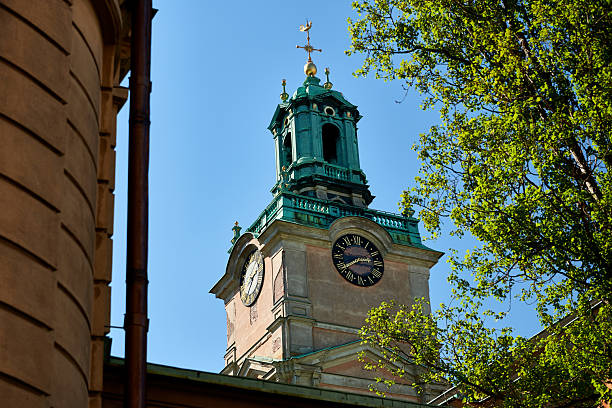 A low-angle, upward-looking view captures the historic clock tower of Storkyrkan (Stockholm Cathedral) against a vibrant blue sky. The tower features a distinctive multi-tiered green copper roof with a patina, topped by a golden finial and weather vane. Two clock faces are visible on the weathered, light-colored stone facade, one prominently displaying Roman numerals. On the right, bright green spring foliage from a tree frames the tower, while a section of an adjacent building and a brown downspout are visible on the left, adding context to the urban setting in Stockholm, Sweden.