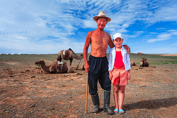 A shirtless older man in a wide-brimmed hat and rubber boots stands with a walking stick, his arm around a young child wearing a white cap and bright summer clothes. Several saddled camels lie on the dry, open steppe behind them, with a few white gers visible on the distant horizon beneath a bright blue, cloud-streaked sky. The image captures a quiet pastoral moment and a warm intergenerational connection.