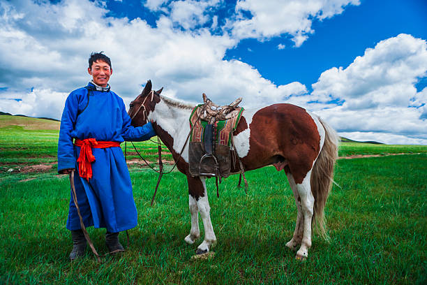 A young herder stands on a wide, green grassland holding the reins of a saddled piebald horse. He wears a bright blue traditional robe tied with a red sash; the horse has a richly patterned saddle blanket and leather tack. Rolling hills stretch into the distance under a vivid blue sky filled with billowing white clouds, creating a peaceful, open landscape scene.
